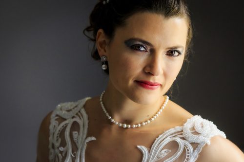 Author: Dedans PhotographyPortrait of the bride against clean gray background, natural light from window, wearing pearl necklace and earrings, looking straight into the camera.